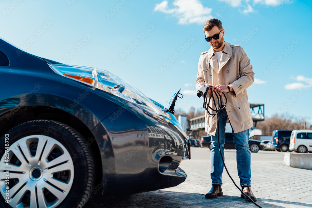 Man with smartphone folds car charger cable after finishing charging of battery. Male near electric vehicle Man with smartphone folds car charger cable after finishing charging of battery. Male near electric vehicle