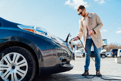 Stylish man in sunglasses inserts the charging cable into the socket of electric car