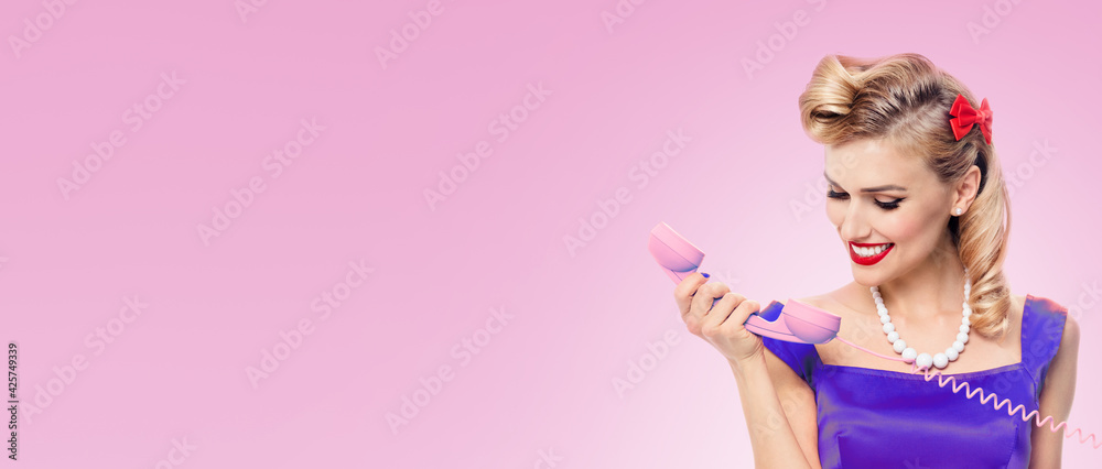 Portrait of beautiful happy smiling woman holding phone tube, dressed in pinup style violet dress, over pink color background. Caucasian blond girl posing in retro studio image.
