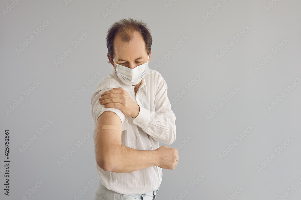 Mature patient in protective face mask recommends coronavirus vaccination. Senior man on gray studio background shows arm with rolled up sleeve and adhesive plaster after receiving Covid 19 vaccine Mature patient in protective face mask recommends coronavirus vaccination. Senior man on gray studio background shows arm with rolled up sleeve and adhesive plaster after receiving Covid 19 vaccine
