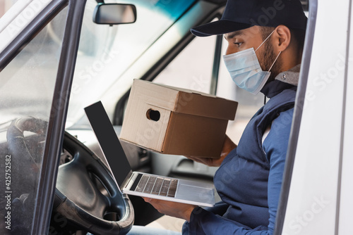 muslim courier in medical mask sitting with laptop in car