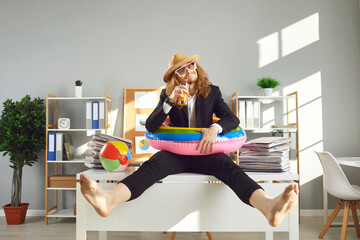 Annual leave and planning travel vacation concept. Funny office worker waiting for summer holidays. Happy barefoot employee in sun hat, sunglasses and swim ring sitting on desk and drinking cocktail