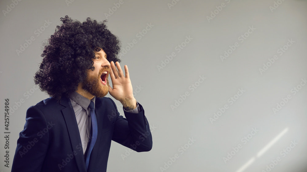 Funny man in curly wig puts hand to open mouth and screams loudly. Crazy office worker in suit yelling standing in profile on gray advertising copy space background. Announcement and sale concept Funny man in curly wig puts hand to open mouth and screams loudly. Crazy office worker in suit yelling standing in profile on gray advertising copy space background. Announcement and sale concept