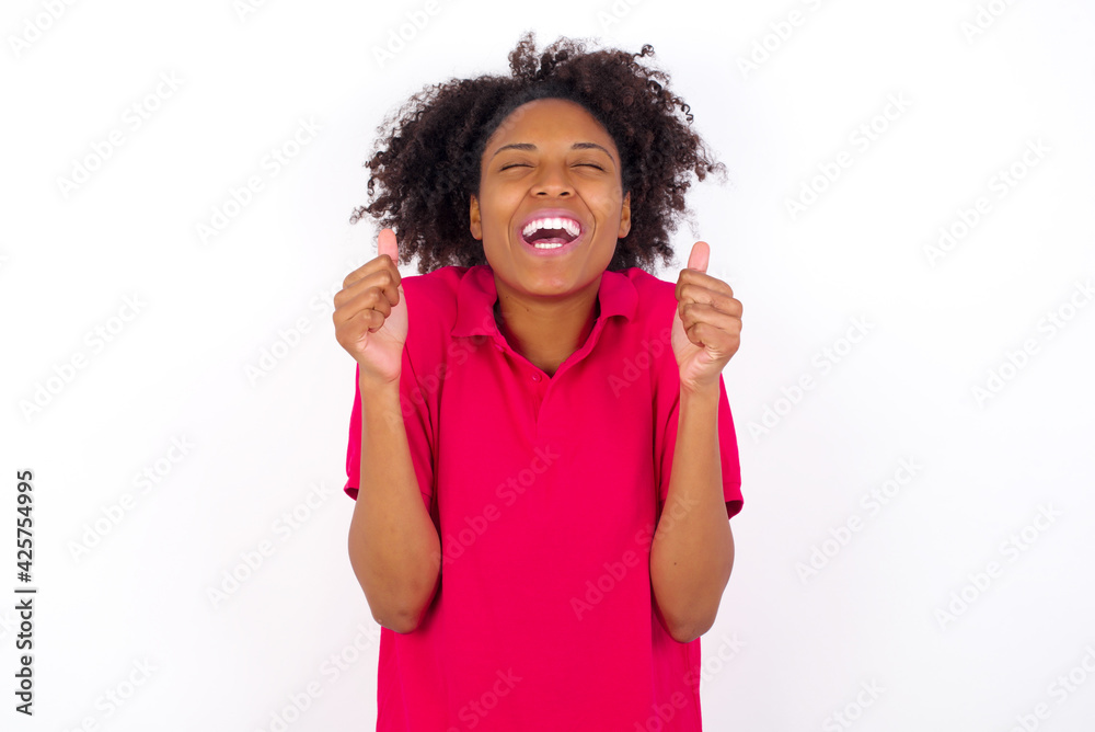 young beautiful African American woman wearing pink t-shirt against white rejoicing his success and victory clenching fists with joy being happy to achieve aim and goals. Positive emotions, feelings. young beautiful African American woman wearing pink t-shirt against white rejoicing his success and victory clenching fists with joy being happy to achieve aim and goals. Positive emotions, feelings.