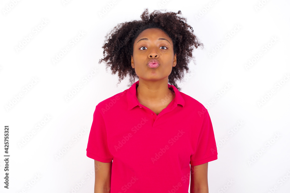 young beautiful African American woman wearing pink t-shirt against white wall , keeps lips as going to kiss someone, has glad expression, grimace face. Standing indoors. Beauty concept. young beautiful African American woman wearing pink t-shirt against white wall , keeps lips as going to kiss someone, has glad expression, grimace face. Standing indoors. Beauty concept.