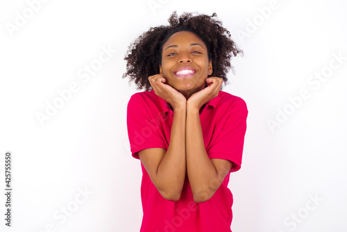 Dreamy young beautiful African American woman wearing pink t-shirt against white wall keeps hands pressed together under chin, looks with happy expression, has toothy smile.