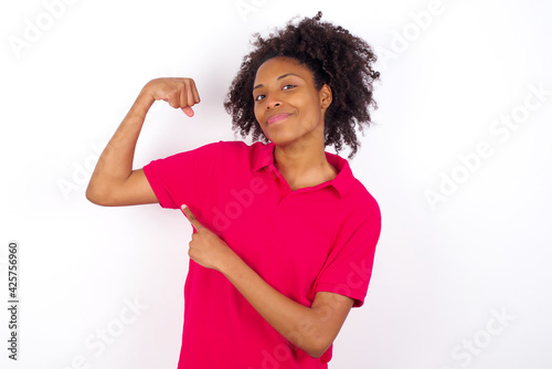 Smiling young beautiful African American woman wearing pink t-shirt against white wall raises hand to show muscles, feels confident in victory, strong and independent.