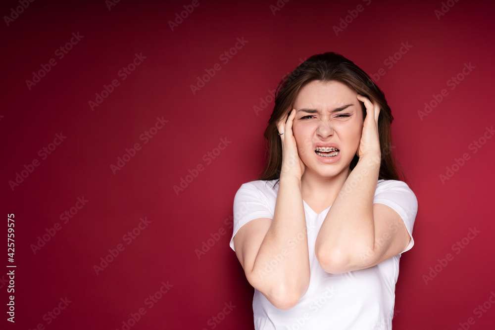A beautiful girl in a white T-shirt in braces holds her head with her hands and twists her mouth from pain in her teeth A beautiful girl in a white T-shirt in braces holds her head with her hands and twists her mouth from pain in her teeth