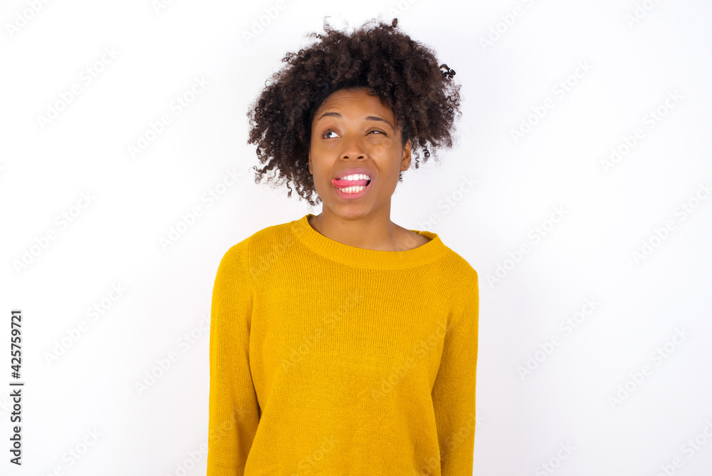 Funny young beautiful African American woman wearing yellow sweater against white wall makes grimace and crosses eyes plays fool has fun alone sticks out tongue. Funny young beautiful African American woman wearing yellow sweater against white wall makes grimace and crosses eyes plays fool has fun alone sticks out tongue.