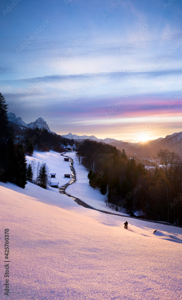 Beautiful pink sunset in the mountains with leading line road towards church and motorbiker having fun in fresh snow