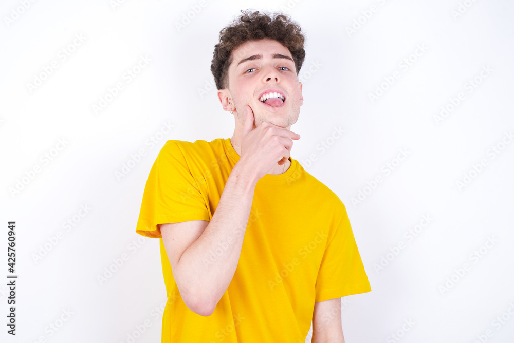 Optimistic young caucasian handsome man with curly hair wearing yellow T-shirt against white keeps hands partly crossed and hand under chin, looks at camera with pleasure. Happy emotions concept. Optimistic young caucasian handsome man with curly hair wearing yellow T-shirt against white keeps hands partly crossed and hand under chin, looks at camera with pleasure. Happy emotions concept.