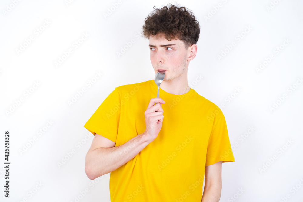 Very hungry young caucasian handsome man with curly hair wearing yellow T-shirt against white studio background holding spoon into mouth dream of tasty meal Very hungry young caucasian handsome man with curly hair wearing yellow T-shirt against white studio background holding spoon into mouth dream of tasty meal