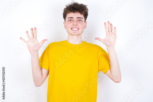 young caucasian handsome man with curly hair wearing yellow T-shirt against white studio background showing and pointing up with fingers number ten while smiling confident and happy.