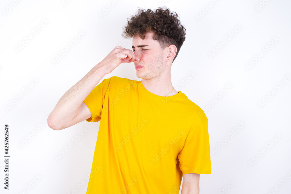 young caucasian handsome man with curly hair wearing yellow T-shirt against white studio smelling something stinky and disgusting, intolerable smell, holding breath with fingers on nose. Bad smell young caucasian handsome man with curly hair wearing yellow T-shirt against white studio smelling something stinky and disgusting, intolerable smell, holding breath with fingers on nose. Bad smell