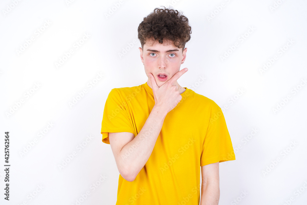young caucasian handsome man with curly hair wearing yellow T-shirt against white studio background Looking fascinated with disbelief, surprise and amazed expression with hands on chin young caucasian handsome man with curly hair wearing yellow T-shirt against white studio background Looking fascinated with disbelief, surprise and amazed expression with hands on chin