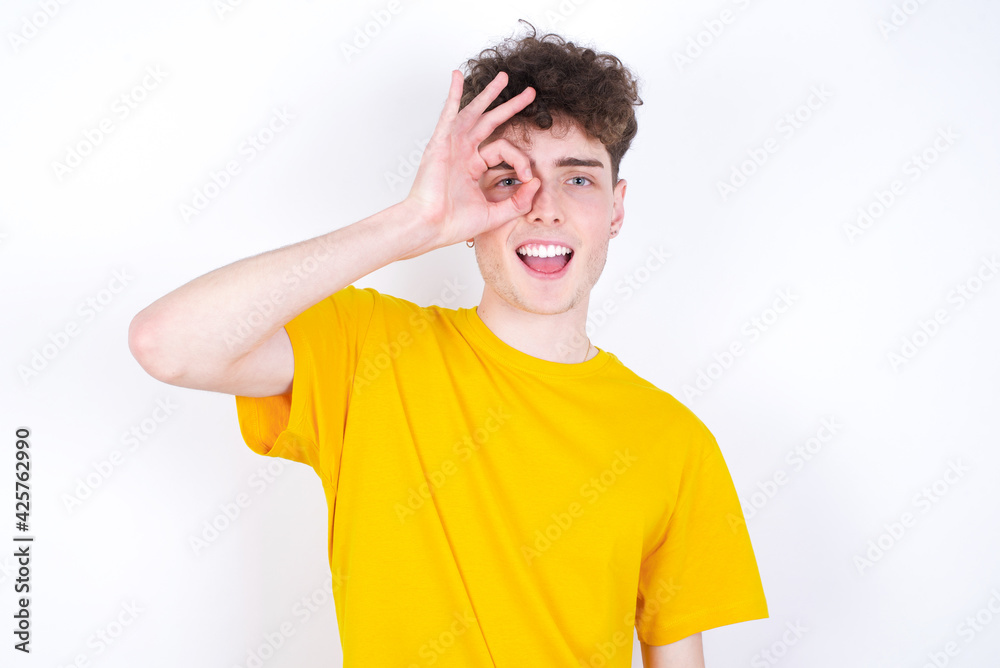 young caucasian handsome man with curly hair wearing yellow T-shirt against white studio background  doing ok gesture with hand smiling, eye looking through fingers with happy face.