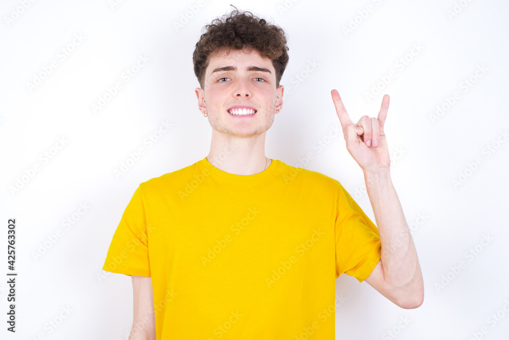 young caucasian handsome man with curly hair wearing yellow T-shirt against white studio background doing a rock gesture and smiling to the camera. Ready to go to her favorite band concert. young caucasian handsome man with curly hair wearing yellow T-shirt against white studio background doing a rock gesture and smiling to the camera. Ready to go to her favorite band concert.