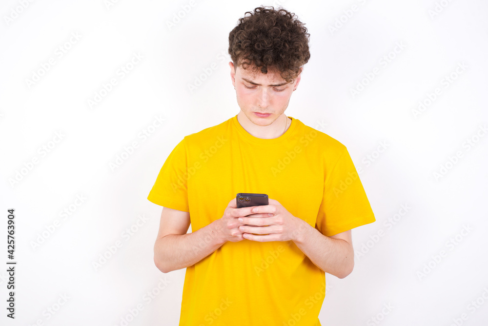 Excited young caucasian handsome man with curly hair wearing yellow T-shirt against white studio background winking and eye hold smart phone use read social network news Excited young caucasian handsome man with curly hair wearing yellow T-shirt against white studio background winking and eye hold smart phone use read social network news