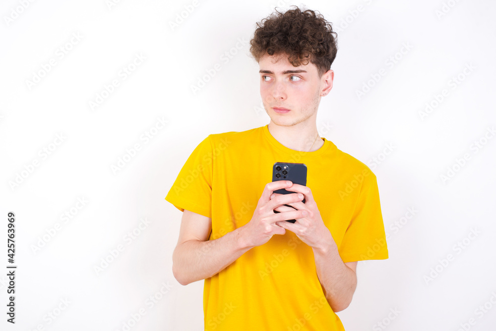 young caucasian handsome man with curly hair wearing yellow T-shirt against white studio background holds telephone hands reads good youth news look empty space advert young caucasian handsome man with curly hair wearing yellow T-shirt against white studio background holds telephone hands reads good youth news look empty space advert
