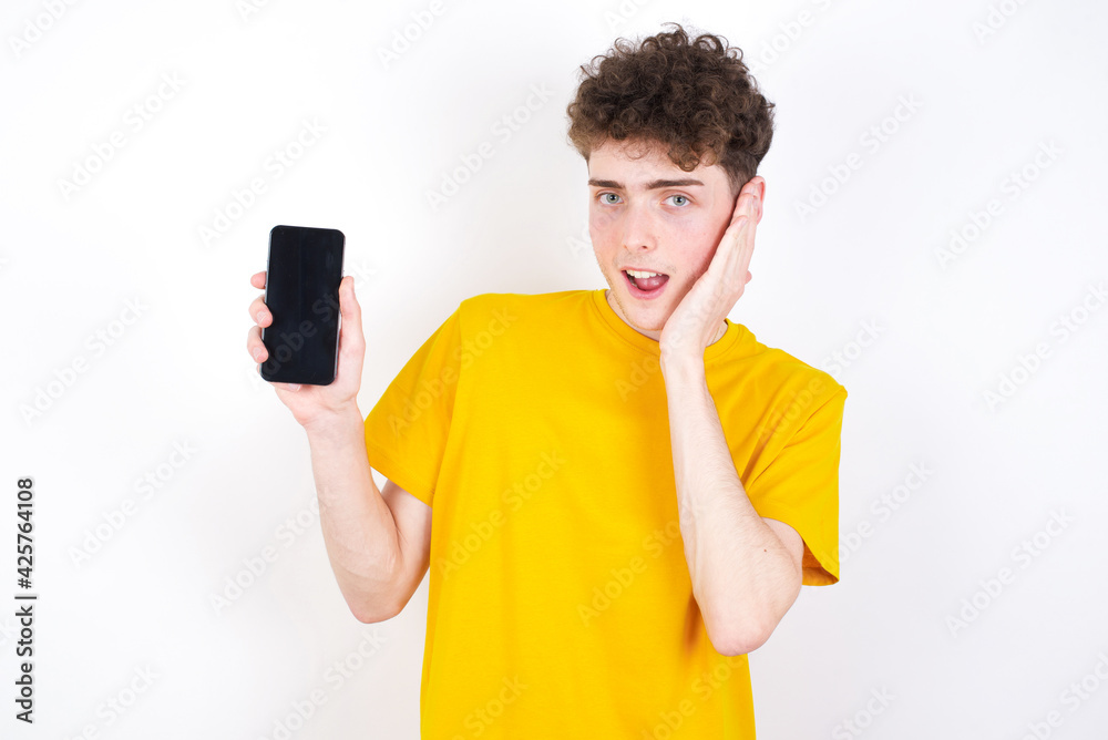 young caucasian handsome man with curly hair wearing yellow T-shirt against white studio hold hand modern technology use touch face palm astonished impressed scream wow omg unbelievable unexpected young caucasian handsome man with curly hair wearing yellow T-shirt against white studio hold hand modern technology use touch face palm astonished impressed scream wow omg unbelievable unexpected