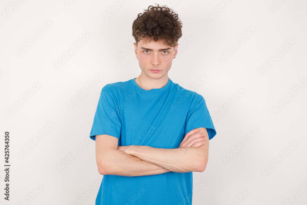 Self confident serious calm young caucasian handsome man with curly hair wearing blue T-shirt against white studio background stands with arms folded. Shows professional vibe stands in assertive pose.