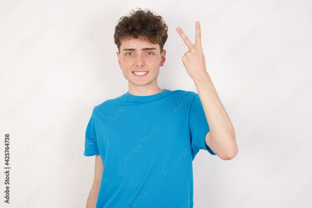 Fototapeta premium young caucasian handsome man with curly hair wearing blue T-shirt against white studio background smiling and looking friendly, showing number two or second with hand forward, counting down