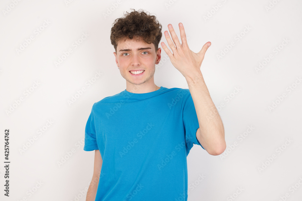 young caucasian handsome man with curly hair wearing blue T-shirt against white studio background smiling and looking friendly, showing number five or fifth with hand forward, counting down young caucasian handsome man with curly hair wearing blue T-shirt against white studio background smiling and looking friendly, showing number five or fifth with hand forward, counting down