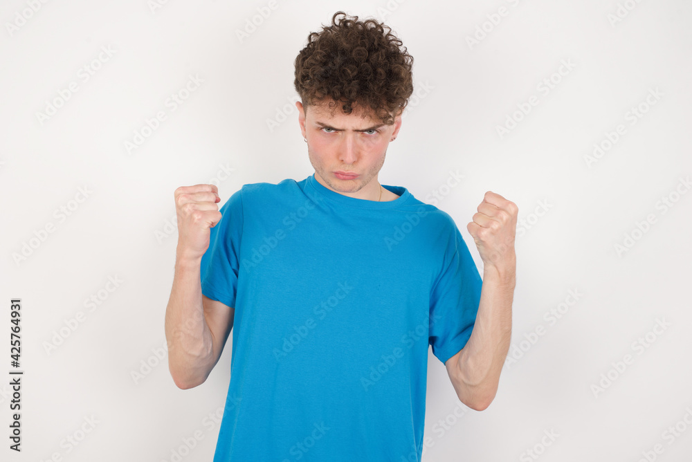 Irritated young caucasian handsome man with curly hair wearing blue T-shirt against white studio blows cheeks with anger and raises clenched fists expresses rage and aggressive emotions. Furious model Irritated young caucasian handsome man with curly hair wearing blue T-shirt against white studio blows cheeks with anger and raises clenched fists expresses rage and aggressive emotions. Furious model