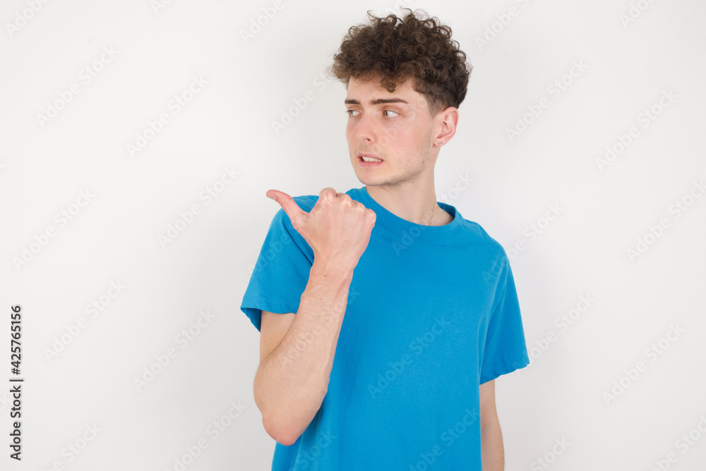 young caucasian handsome man with curly hair wearing blue T-shirt against white studio background points away and gives advice demonstrates advertisement