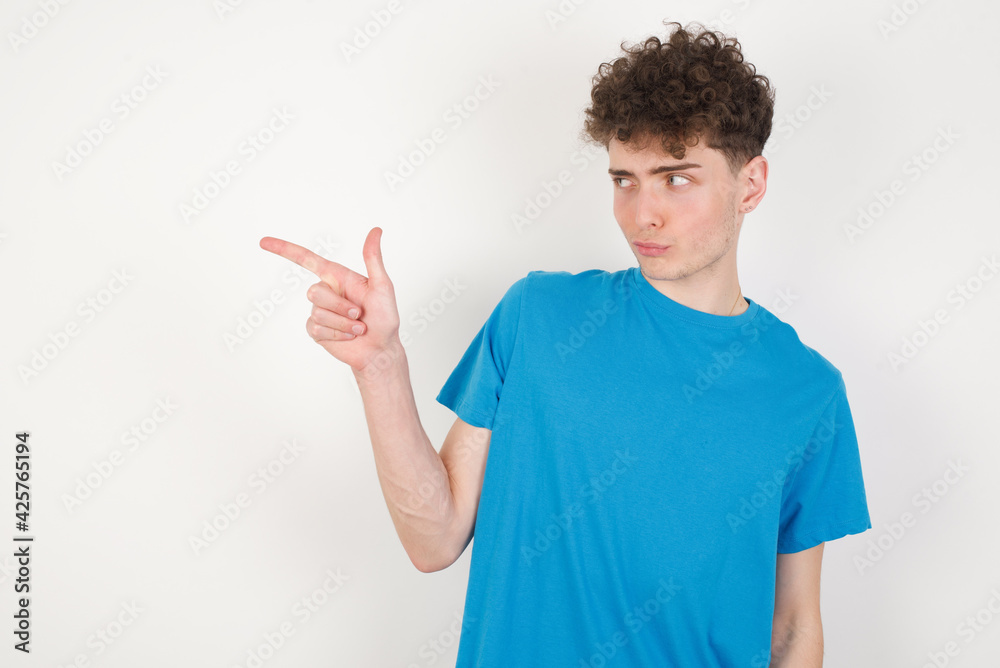 young caucasian handsome man with curly hair wearing blue T-shirt against white studio background points at copy space and advertises something, advices best price. young caucasian handsome man with curly hair wearing blue T-shirt against white studio background points at copy space and advertises something, advices best price.
