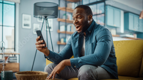 Excited Black African American Man Having a Video Call on Smartphone while Sitting on a Sofa in Living Room. Happy Man Smiling at Home and Talking to His Friends and Family Over the Internet.