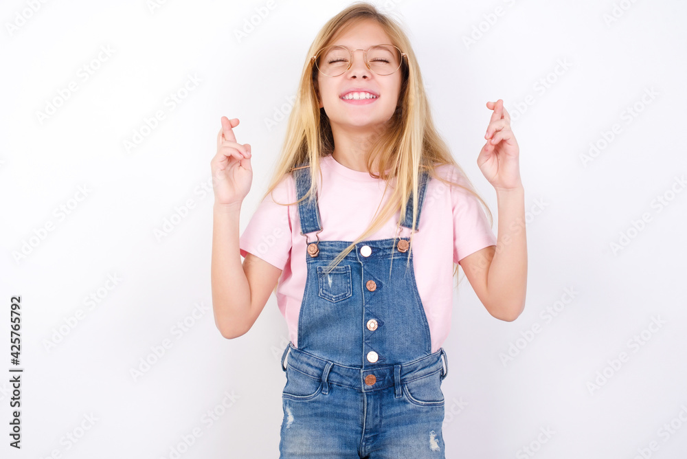 beautiful caucasian little girl wearing jeans overall over white background has big hope, crosses fingers, believes in good fortune, smiles broadly. People and wish concept beautiful caucasian little girl wearing jeans overall over white background has big hope, crosses fingers, believes in good fortune, smiles broadly. People and wish concept