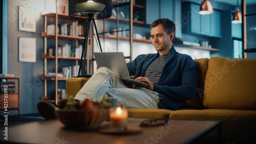 Handsome Caucasian Man Working on Laptop Computer while Sitting on a Sofa in Stylish Cozy Living Room. Freelancer Working From Home. Browsing Internet, Using Social Networks, Having Fun in Flat. photo