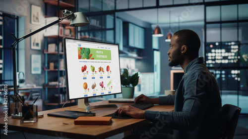 Handsome Black African American is Using Desktop Computer with Groceries Delivery Web Page to Order Fresh Vegetables to Make Dinner. He Lives in Modern Huge Appatment.