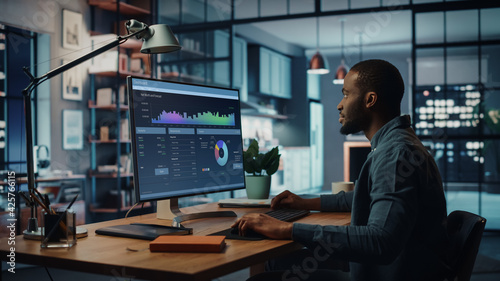 Handsome Black African American Specialist Working on Desktop Computer in Creative Home Living Room. Freelance Male is Doing Market Analysis and Creates Report with Charts for Clients and Employer.