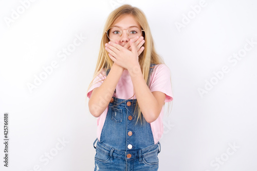 beautiful caucasian little girl wearing denim jeans overall over white background shocked covering mouth with hands for mistake. Secret concept.