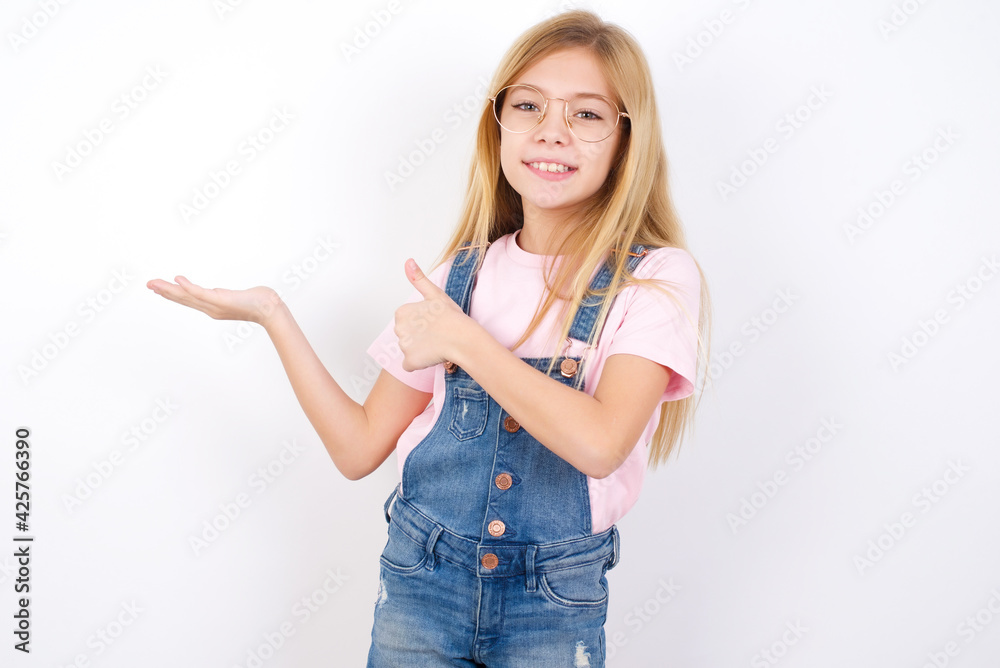 Obraz premium beautiful caucasian little girl wearing denim jeans overall over white background Showing palm hand and doing ok gesture with thumbs up, smiling happy and cheerful.