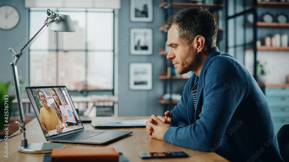 Handsome Caucasian Man Having a Video Call on Laptop Computer while Sitting Behind Desk in Living Room. Freelancer Working From Home and Talking to Colleagues and Clients Over the Internet.