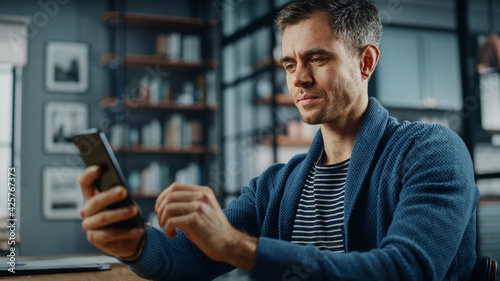 Serious Caucasian Man Using Smartphone while Sitting at a Desk in Stylish Living Room. Young Man Smiling at Home and Chatting to Colleagues and Clients Over the Internet. Using Social Networks.