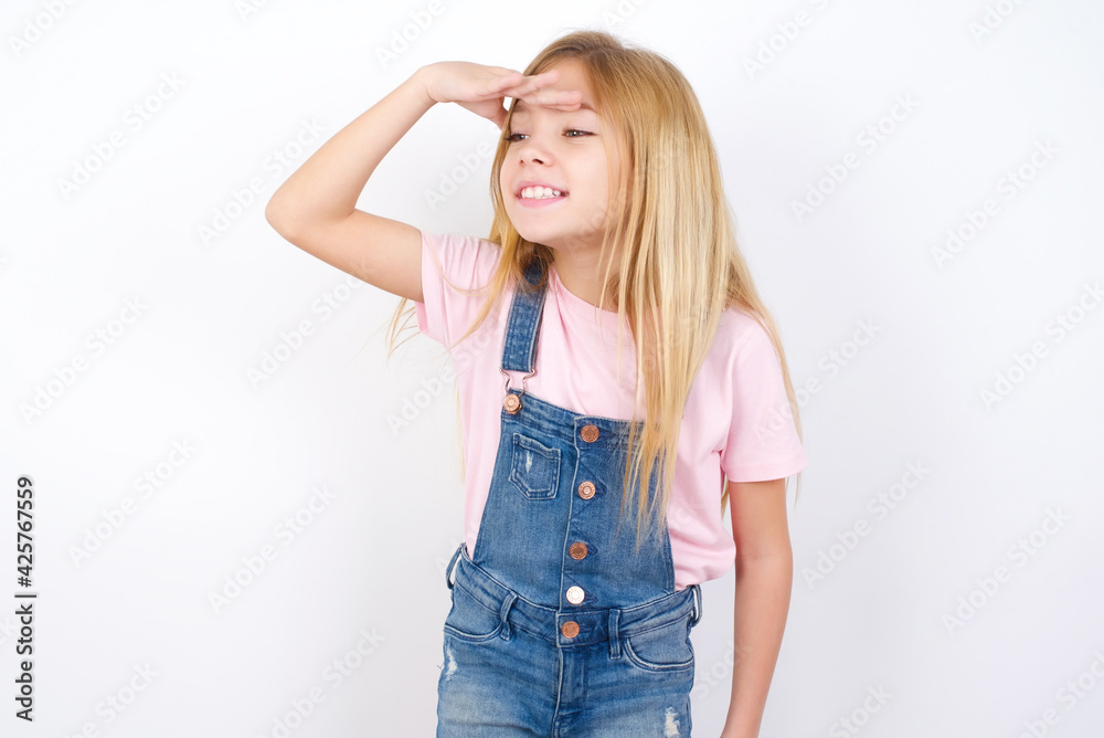 beautiful caucasian little girl wearing denim jeans overall over white background very happy and smiling looking far away with hand over head. Searching concept. beautiful caucasian little girl wearing denim jeans overall over white background very happy and smiling looking far away with hand over head. Searching concept.
