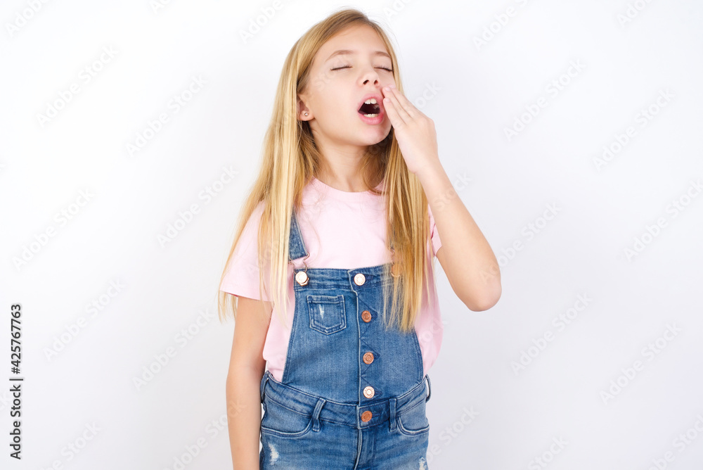 beautiful caucasian little girl wearing denim jeans overall over white background being tired and yawning after spending all day at work. beautiful caucasian little girl wearing denim jeans overall over white background being tired and yawning after spending all day at work.