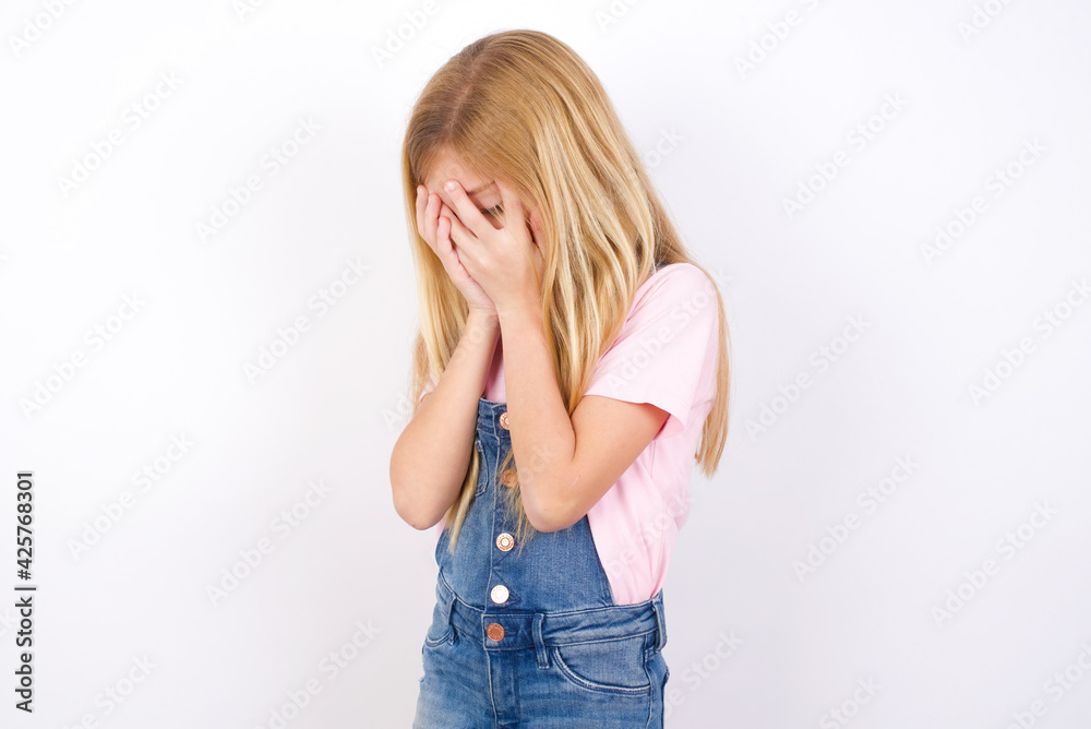 Sad beautiful caucasian little girl wearing denim jeans overall over white background crying covering her face with her hands. Sad beautiful caucasian little girl wearing denim jeans overall over white background crying covering her face with her hands.