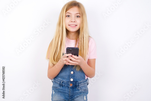 Excited beautiful caucasian little girl wearing denim jeans overall over white background holding smartphone and looking amazed to the camera after receiving good news.
