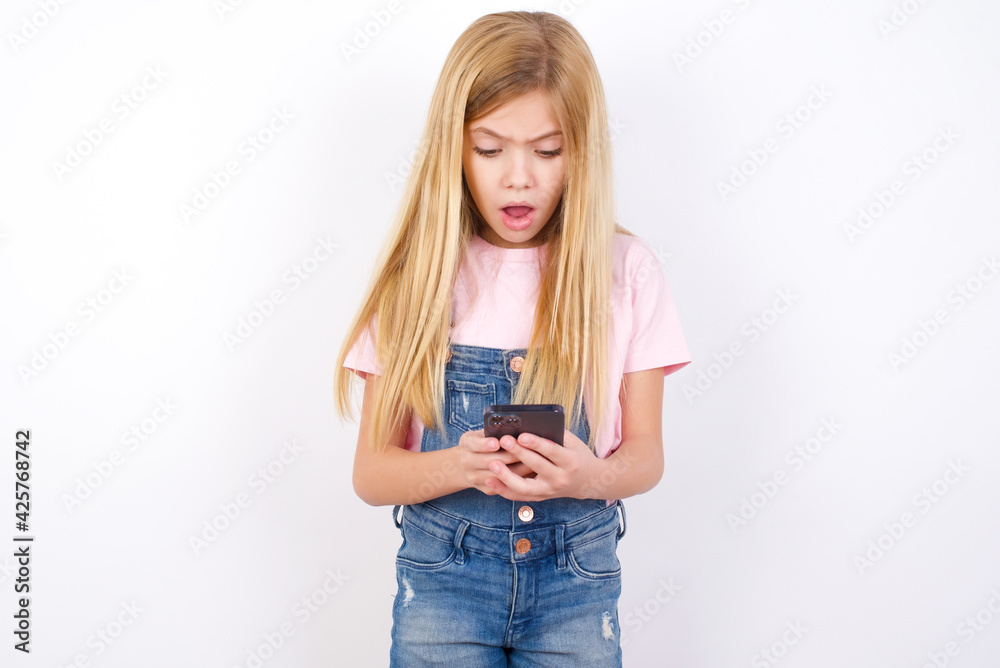 Focused beautiful caucasian little girl wearing denim jeans overall over white background use smartphone reading social media news, or important e-mail