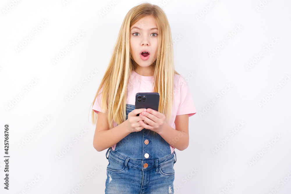 Shocked beautiful caucasian little girl wearing denim jeans overall over  white background opens mouth hold phone reading advert unbelievable big  shopping prices Photos | Adobe Stock