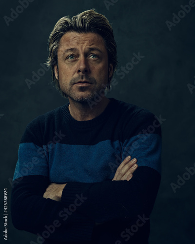 Blond pensive man with arms crossed and stubble beard in blue striped wool sweater in front of a dark wall. Looking towards the camera.