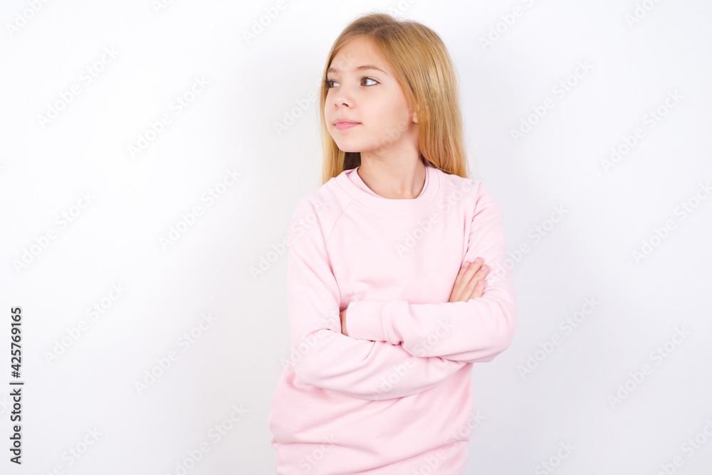 Charming thoughtful Beautiful caucasian blonde little girl wearing pink sweater stands with arms folded concentrated somewhere with pensive expression thinks what to do Charming thoughtful Beautiful caucasian blonde little girl wearing pink sweater stands with arms folded concentrated somewhere with pensive expression thinks what to do