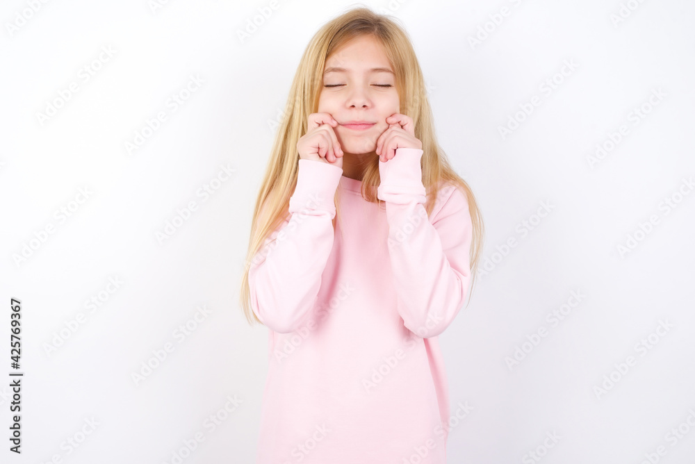 Pleased Beautiful caucasian blonde little girl wearing pink sweater with closed eyes keeps hands near cheeks and smiles tenderly imagines something very pleasant Pleased Beautiful caucasian blonde little girl wearing pink sweater with closed eyes keeps hands near cheeks and smiles tenderly imagines something very pleasant