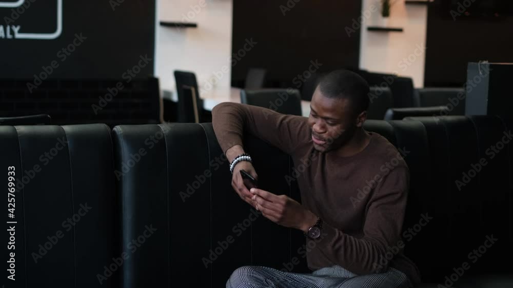 A beautiful young and happy African man uses a smartphone while sitting in a cafe with a dark interior. He is browsing the Internet or social networks while waiting for friends or departure