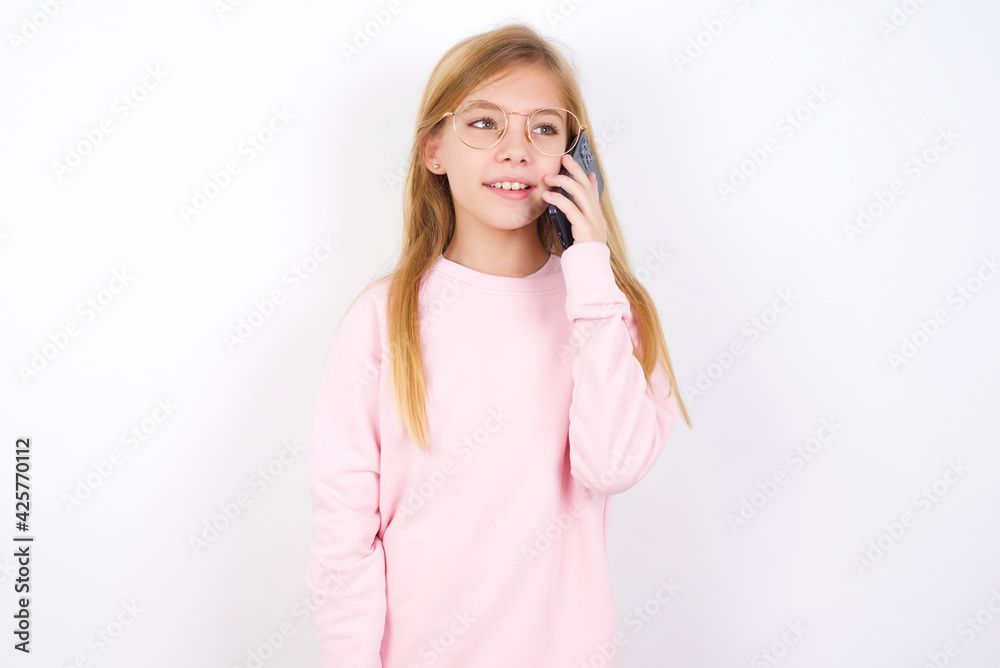 Pleasant looking happy beautiful caucasian little girl wearing pink sweater has nice telephone conversation and looks aside, has nice mood and smiles positively while talks via cell phone Pleasant looking happy beautiful caucasian little girl wearing pink sweater has nice telephone conversation and looks aside, has nice mood and smiles positively while talks via cell phone